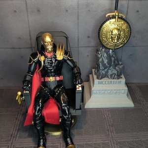 Può includere: Un action figure calvo con testa dorata, che indossa un'armatura nera con dettagli dorati e un mantello rosso, seduto su una sedia nera. Accanto c'è uno scudo dorato con un volto e una spada, montato su una base grigia con la scritta MCCULLEN.
