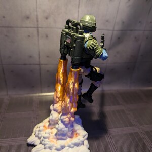 Pu&ograve; includere: Una action figure verde e nera con uno zaino a razzo e fiamme arancioni. La figura indossa un casco e sta in piedi su una nuvola bianca con fiamme arancioni.