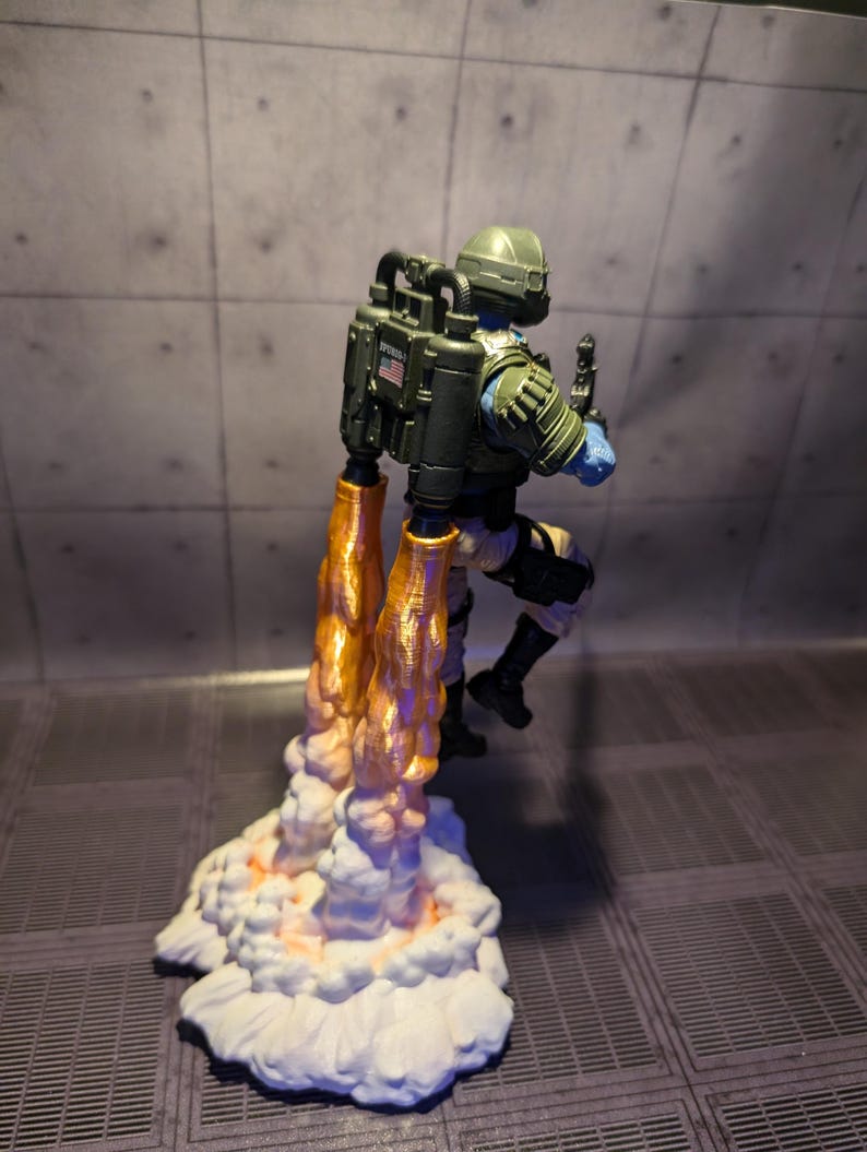 Pu&ograve; includere: Una action figure verde e nera con uno zaino a razzo e fiamme arancioni. La figura indossa un casco e sta in piedi su una nuvola bianca con fiamme arancioni.