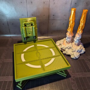 Pu&ograve; includere: Una piattaforma di atterraggio verde con un cerchio bianco al centro. Un jetpack giocattolo verde e nero &egrave; attaccato al muro dietro la piattaforma di atterraggio. Due scarichi di razzi giocattolo arancioni e bianchi sono sul lato destro dell'immagine.