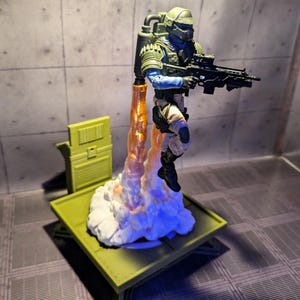 Pu&ograve; includere: Una action figure di un soldato con un elmetto e un fucile. La figura &egrave; in piedi su una piattaforma con una nuvola bianca e fiamme arancioni che escono dal fondo.