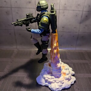 May include: A green and black action figure with a jetpack and a gun, standing on a white cloud with an orange flame coming out of the jetpack.