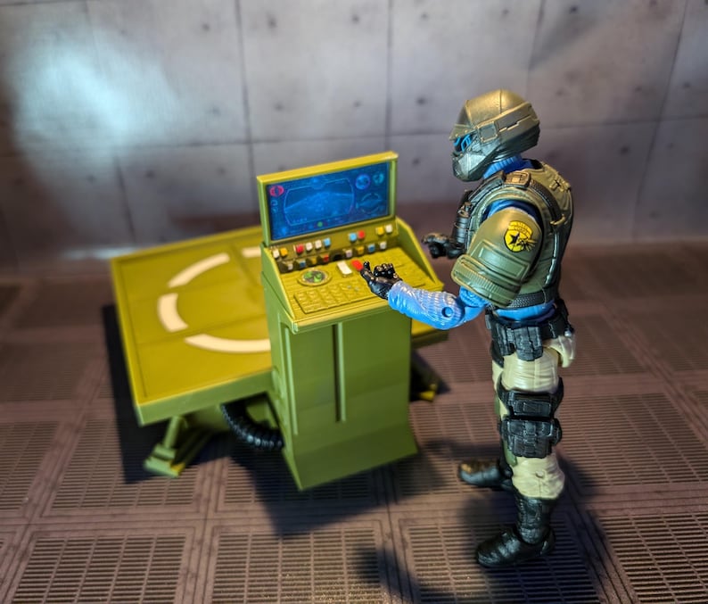 Pu&ograve; includere: Un pannello di controllo verde e grigio con uno schermo che mostra un'immagine radar. Una piattaforma verde e grigia con un design circolare &egrave; sullo sfondo. Una action figure verde e grigia con un casco &egrave; in piedi accanto al pannello di controllo.