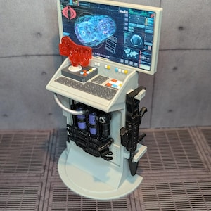 May include: A gray and black plastic toy computer console with a red button and a screen displaying a blue and white graphic. The console has a keyboard, a monitor, and a variety of weapons and accessories attached to the sides.