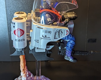 May include: A white action figure jetpack with a clear plastic canopy, a blue and purple figure inside, and a pink and orange smoke effect coming from the bottom. The jetpack has the text "PE 49-LTA" and "15 E5 MQ" on the side.