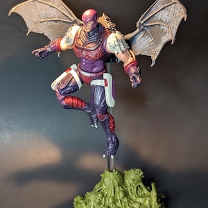 May include: A purple and red action figure with gray wings, standing on a green base. The figure has a purple and white suit with a red belt and a gray mask.