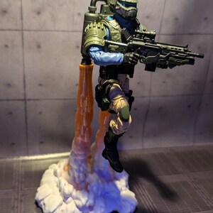 May include: A green and blue action figure of a soldier wearing a helmet and holding a gun. The figure is standing on a white cloud-like base with orange flames coming out of the bottom.
