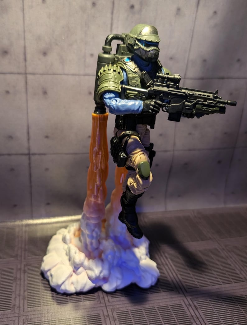 May include: A green and blue action figure of a soldier wearing a helmet and holding a gun. The figure is standing on a white cloud-like base with orange flames coming out of the bottom.