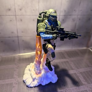 May include: A green and blue action figure of a soldier wearing a helmet and holding a gun. The figure is standing on a white cloud with an orange flame coming out of the bottom.