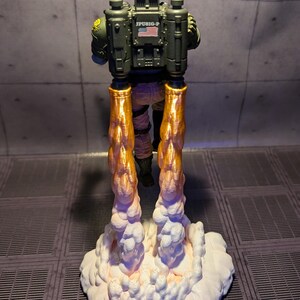 May include: A green and black action figure with a jetpack and a US flag on the back. The figure is flying with orange flames coming out of the jetpack and white smoke below.