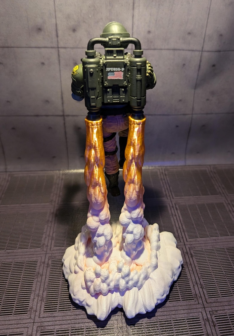 May include: A green and black action figure with a jetpack and a US flag on the back. The figure is flying with orange flames coming out of the jetpack and white smoke below.