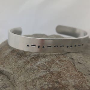 May include: A silver bracelet with a pattern of dots and dashes engraved on the front. The bracelet is resting on a grey rock.