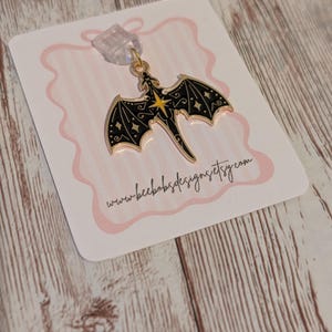 May include: A black and gold dragon charm with detailed wings and a star design. The charm hangs from a clear clip on a pink-edged card. The website address www.beebobsdesigns.etsy.com is printed on the card, set against a wooden background.