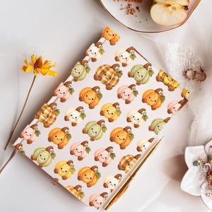 May include: A rectangular notebook with a white cover featuring a repeating pattern of small, smiling cartoon pumpkins in shades of orange, green, pink, and yellow, with some pumpkins displaying a plaid pattern and others in solid colours.