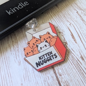 May include: A Kitten Nuggets charm showing a box of cartoon kittens in orange and white. The charm has a red border and the words "Kitten Nuggets". The charm is attached to a black Kindle.
