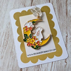 May include: A gold-coloured earring depicting a white rabbit on a yellow crescent moon, adorned with orange and yellow flowers and green leaves. The earring is on a white card with a scalloped gold border. The website address is visible on the card.