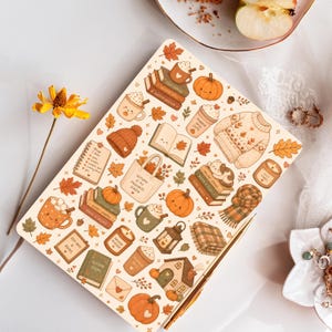 May include: A rectangular notebook featuring a cream-coloured cover with a repeating pattern of autumn-themed illustrations including pumpkins, mugs of hot drinks, books, sweaters, candles, and leaves in shades of orange, brown, and green.