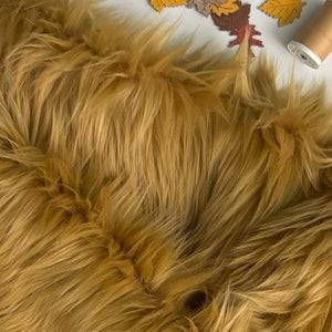 May include: Close-up of a pile of golden-brown faux fur fabric. The fabric has long, shaggy fibers, creating a textured appearance. Decorative fall leaves and a spool of thread are visible in the background.