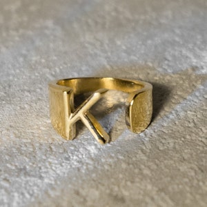 Può includere: Un anello dorato con la lettera "K" in un carattere semplice e moderno.