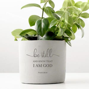 May include: A small potted plant with vibrant green leaves. The plant is in a light gray pot with the words "be still AND KNOW THAT I AM GOD" and the scripture reference Psalms 46:10 in black script. The pot sits on a white surface.