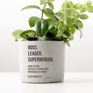 Peut inclure: Un petit pot de fleurs en béton gris clair avec les mots "BOSS. LEADER. SUPERWOMAN." en texte noir. Le pot est rempli de plantes vertes à feuilles. Le texte continue avec "HARD TO FIND, DIFFICULT TO PART WITH, IMPOSSIBLE TO FORGET, ALSO KNOWN AS:"