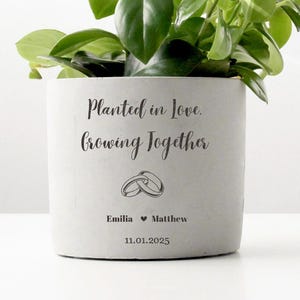 May include: A small, light gray planter pot with a green plant. The pot has the text "Planted in love, growing together" and the names Emilia and Matthew with a date. The pot is on a white surface.