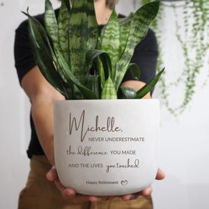 May include: A white, square-shaped plant pot holding a green plant. The pot is inscribed with "Michelle, Never Underestimate the difference YOU MADE AND THE LIVES you touched Happy Retirement". The plant has long, green leaves.