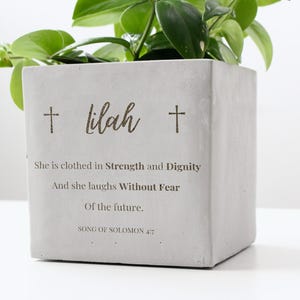 May include: A square, light gray concrete planter with the name "Lilah" and two cross symbols. The planter has text that reads "She is clothed in Strength and Dignity" and "And she laughs Without Fear". Green plant stems and leaves are visible.