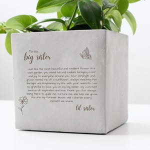May include: A square, light gray concrete planter with a plant inside. The planter has the text "To my big sister" and a poem with a butterfly and flower illustration. The text is in a handwritten style.