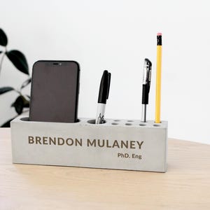 May include: A gray concrete desk organizer with a phone, pen, pencil, and multiple small holes for pens. The text "BRENDON MULANEY, PhD. Eng" is printed on the front. The organizer is on a light wood surface.