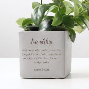 Peut inclure: Un pot de fleurs carré gris clair avec le mot "Friendship" et une citation sur l'amitié imprimée sur le devant. Le pot contient une plante verte à feuilles. Le texte comprend également les noms "Emma & Kylie".