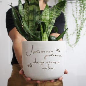 May include: A white, cylindrical planter pot with a snake plant. The pot has the text "Aunts are like gardeners, always nurturing us with love." The snake plant has green and yellow striped leaves. The pot is held by a person.