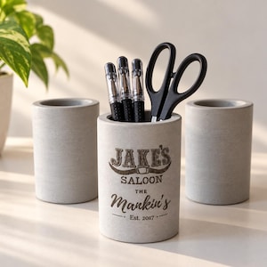 May include: Three cylindrical grey concrete pencil holders. The centre holder features dark text reading JAKE'S SALOON THE Mankin's Est. 2017 with a graphic of a belt buckle. The centre holder contains three black pens and a pair of black scissors.