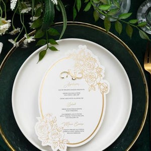May include: A white oval-shaped wedding menu card with gold floral illustrations and text. The menu card is on a white plate with a gold rim, which is on a green tablecloth. The text on the card reads "Bacon Wrapped Water Chestnuts, Spring Egg Rolls with Thai Sweet Chili Sauce, Crab Cakes with Cilantro Aioli, Salad, Fresh Roasted Peppers with Tomato, Basil, and Vinaigrette, Mac & Cheese, Chicken Breast Sauteed in Marsala Wine Topped with Melted Mozzarella and served with Roasted Potatoes".