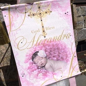 May include: A pink and white banner with a gold cross and the text "God Bless Alexandra". A baby girl is sleeping on a pink blanket.
