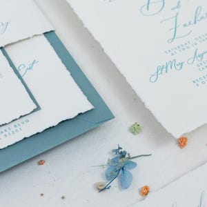 May include: A wedding invitation suite with a blue and white color scheme. The invitation is printed on textured paper with a torn edge. The text on the invitation reads "Saturday, October at two in the afternoon, St. Mary's Church, 5955 Lindle, Tarzana, CA, Reception 7 o'clock, 1320"