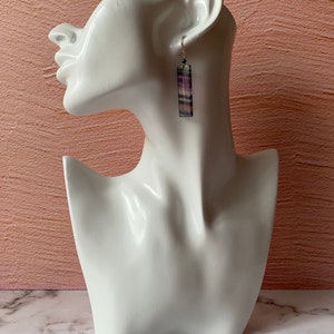 Pode incluir: Brincos pendentes listrados em roxo e branco com um gancho prateado e um detalhe de contas pretas.