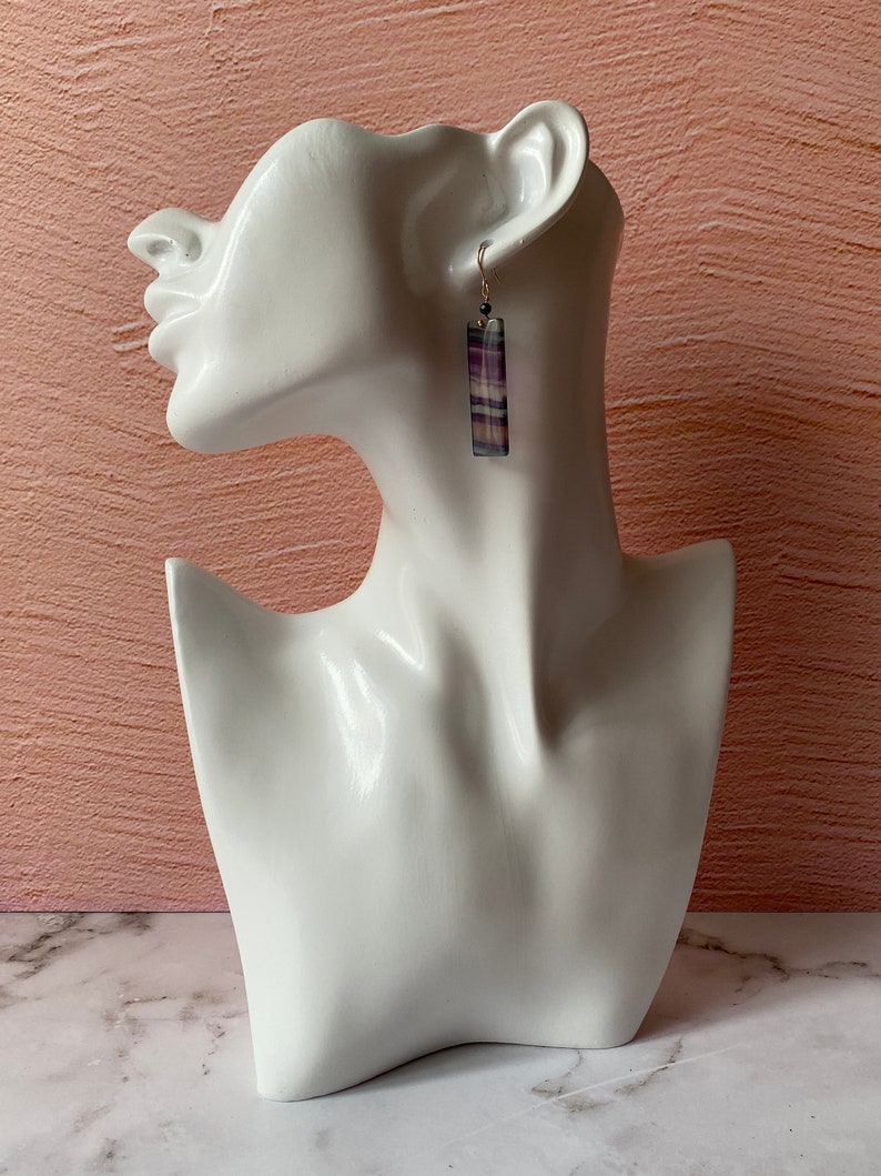 Pode incluir: Brincos pendentes listrados em roxo e branco com um gancho prateado e um detalhe de contas pretas.