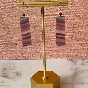 Pode incluir: Um par de brincos de ouro com gotas de fluorita listradas de roxo e branco e contas pretas. Os brincos s&atilde;o exibidos em um suporte de ouro.