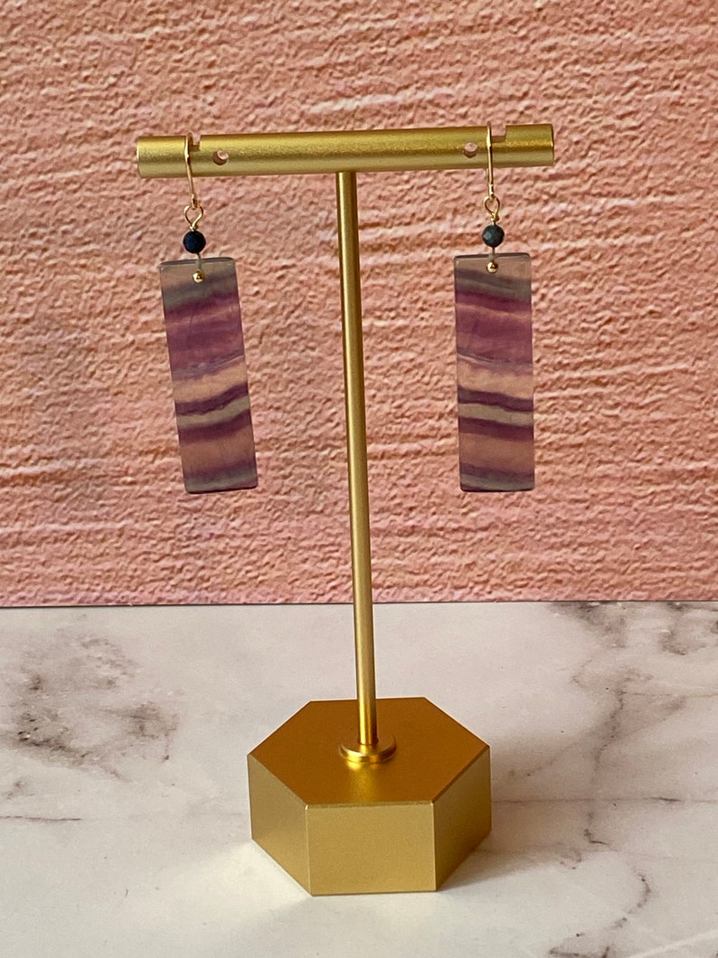 Pode incluir: Um par de brincos de ouro com gotas de fluorita listradas de roxo e branco e contas pretas. Os brincos s&atilde;o exibidos em um suporte de ouro.