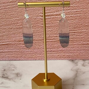 Pode incluir: Um par de brincos de prata com uma pedra de fluorita oval transparente com listras roxas e azuis. Os brincos est&atilde;o pendurados em um suporte dourado.
