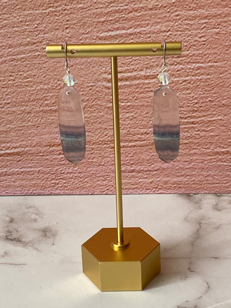 Pode incluir: Um par de brincos de prata com uma pedra de fluorita oval transparente com listras roxas e azuis. Os brincos est&atilde;o pendurados em um suporte dourado.