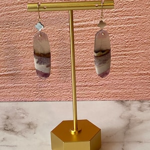 Pode incluir: Um par de brincos de ouro com gotas de pedra de &aacute;gata roxa e branca. Os brincos s&atilde;o exibidos em um suporte de ouro.