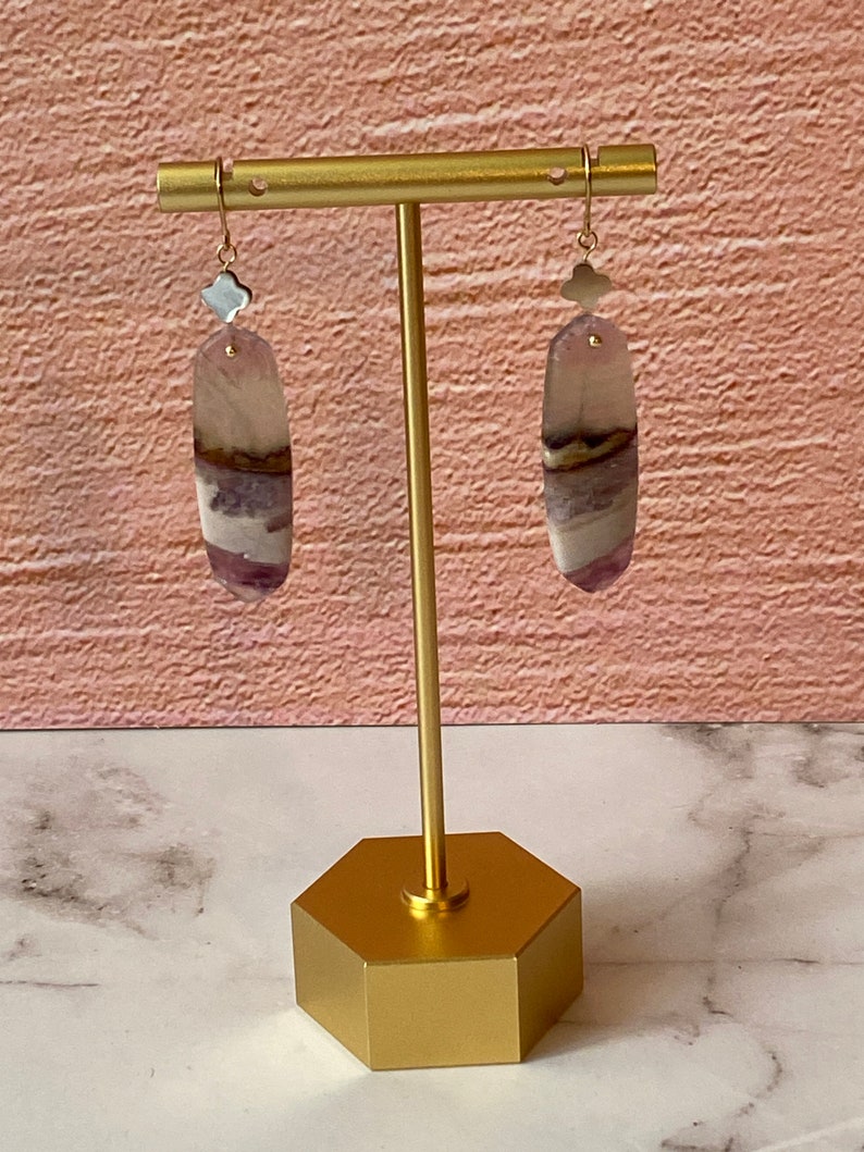 Pode incluir: Um par de brincos de ouro com gotas de pedra de &aacute;gata roxa e branca. Os brincos s&atilde;o exibidos em um suporte de ouro.