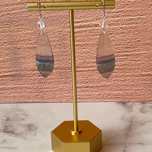 Pode incluir: Um par de brincos pendentes com uma pedra de fluorita em forma de l&aacute;grima em tons de roxo, azul e verde. As pedras s&atilde;o suspensas em ganchos prateados com uma conta de cristal transparente no topo.