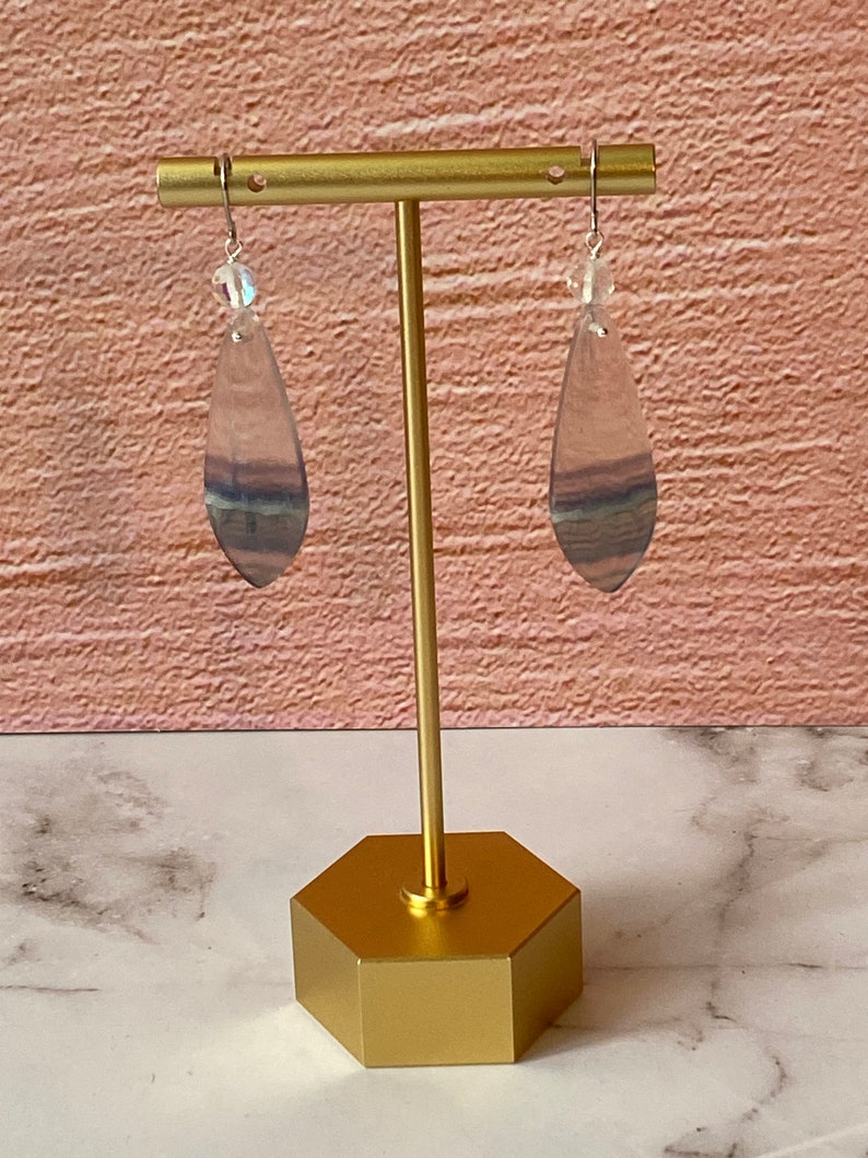 Pode incluir: Um par de brincos pendentes com uma pedra de fluorita em forma de l&aacute;grima em tons de roxo, azul e verde. As pedras s&atilde;o suspensas em ganchos prateados com uma conta de cristal transparente no topo.