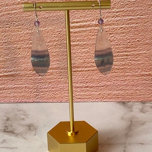 Pode incluir: Um par de brincos pendentes com pedras de fluorita em forma de l&aacute;grima. As pedras s&atilde;o transl&uacute;cidas e t&ecirc;m um padr&atilde;o de cor roxo, verde e branco. Os brincos est&atilde;o pendurados em um suporte dourado.