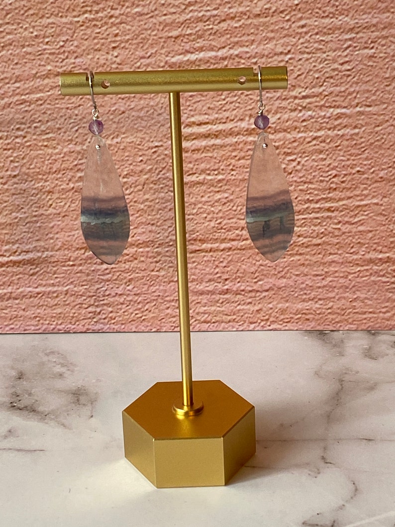 Pode incluir: Um par de brincos pendentes com pedras de fluorita em forma de l&aacute;grima. As pedras s&atilde;o transl&uacute;cidas e t&ecirc;m um padr&atilde;o de cor roxo, verde e branco. Os brincos est&atilde;o pendurados em um suporte dourado.