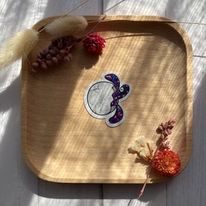 May include: A purple and white glitter sticker of a turtle biting into a moon. The sticker is on a wooden tray with dried flowers.