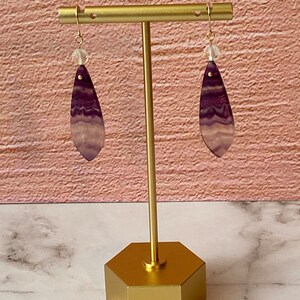 Pode incluir: Um par de brincos pendentes com pedras em forma de l&aacute;grima listradas de roxo e branco e contas de cristal transparentes penduradas em ganchos dourados.